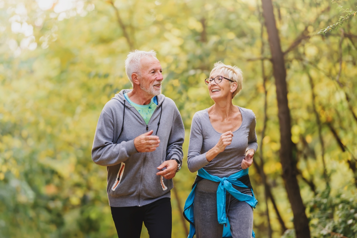 Couple de seniors faisant du jogging ensemble dans un parc boisé, souriants et en tenue de sport.
