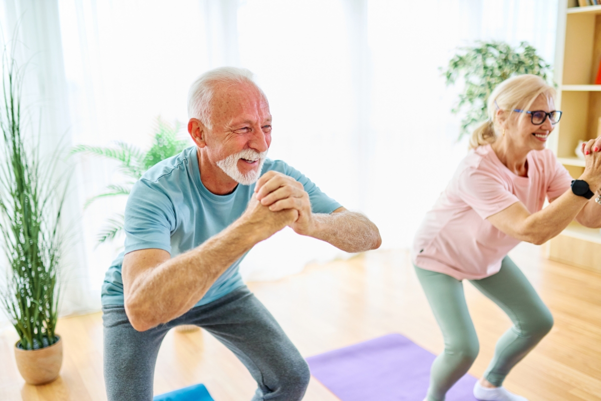 Homme et femme seniors effectuant des exercices de renforcement musculaire (squats) chez eux, sans équipement.