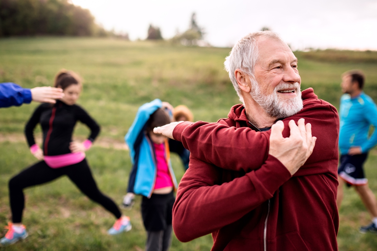 Homme senior s’étirant en plein air pendant une séance de fitness en groupe, avec d’autres participants en arrière-plan. 