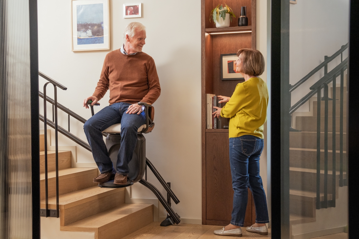 Un homme âgé utilise un monte-escalier dans une maison moderne pendant qu’une femme se tient à proximité. 