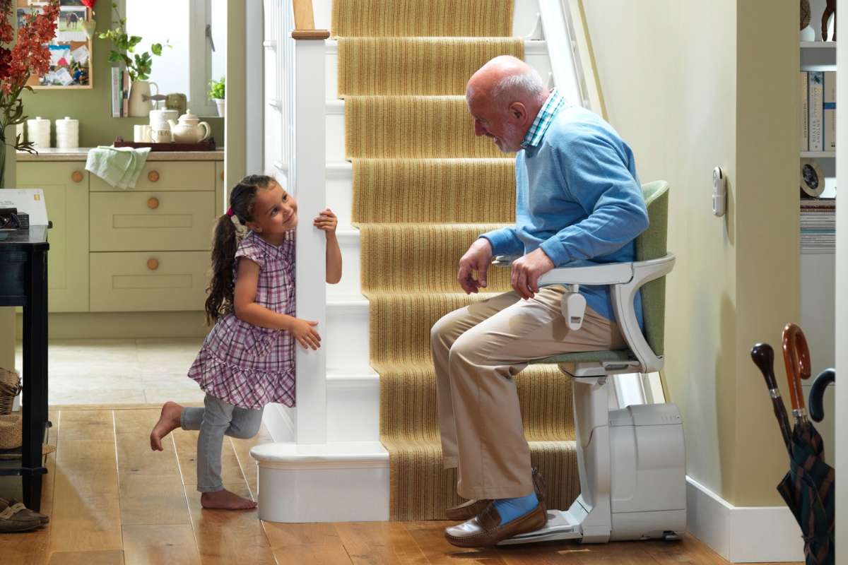Un homme âgé assis sur un monte-escalier sourit et discute avec une petite fille qui regarde autour de l’escalier dans une maison chaleureuse. 