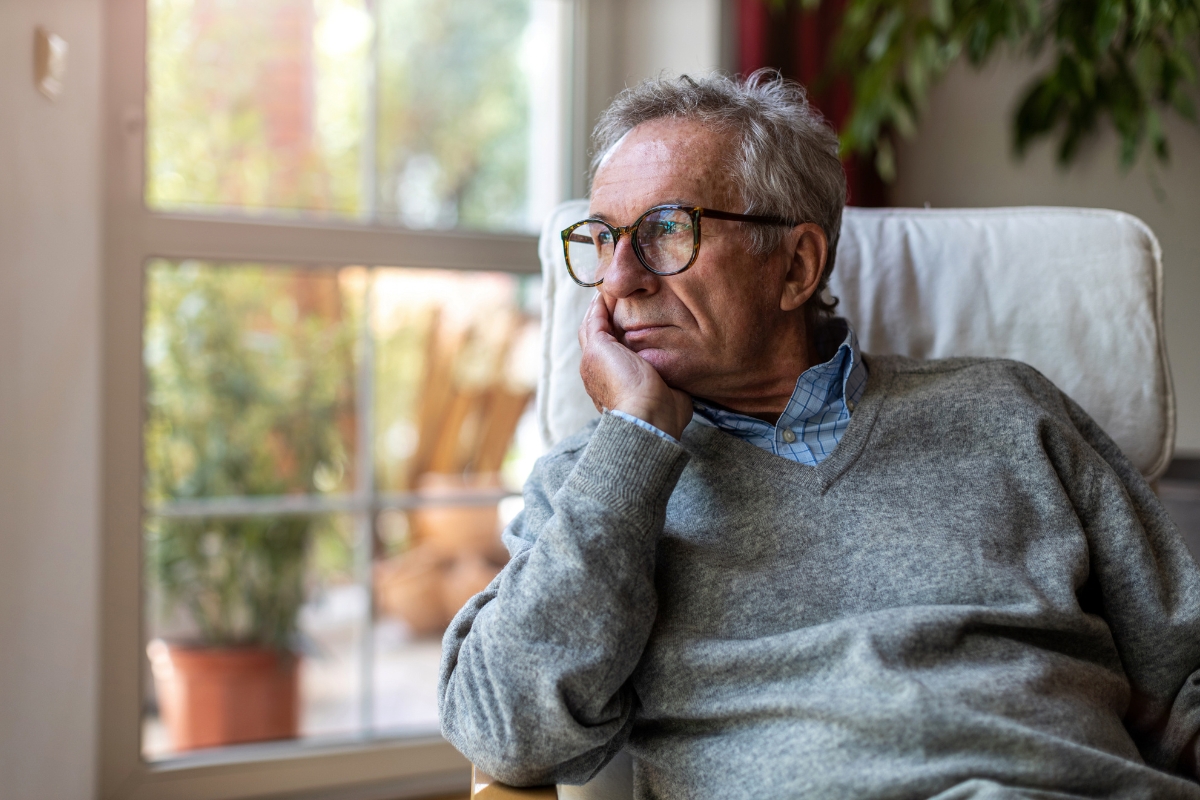 Un homme âgé aux cheveux gris et portant des lunettes est assis près d’une grande fenêtre, la tête appuyée sur sa main, regardant dehors d’un air pensif.
