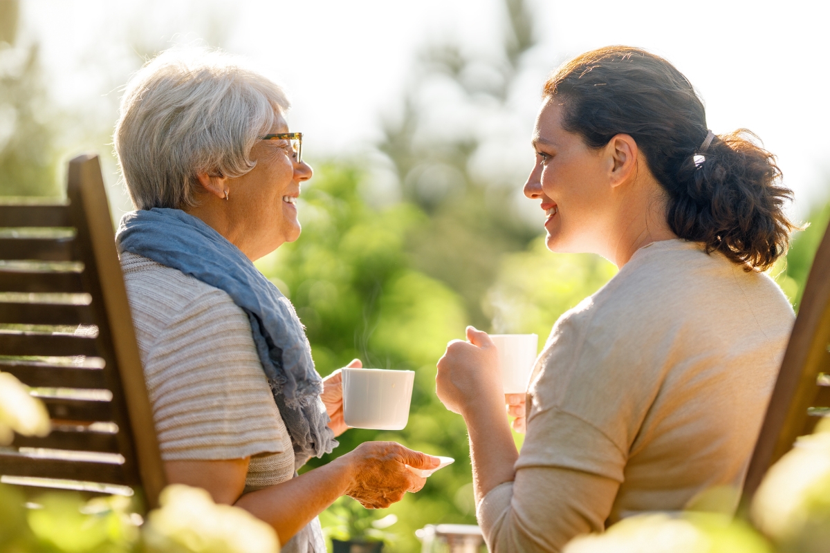 Une femme âgée et une femme plus jeune sont assises dehors avec des tasses à la main, souriant pendant une conversation détendue dans un jardin ensoleillé.