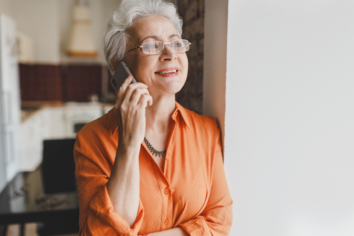 Une femme âgée souriante aux cheveux blancs et portant des lunettes parle au téléphone portable à l’intérieur, près d’une fenêtre.
