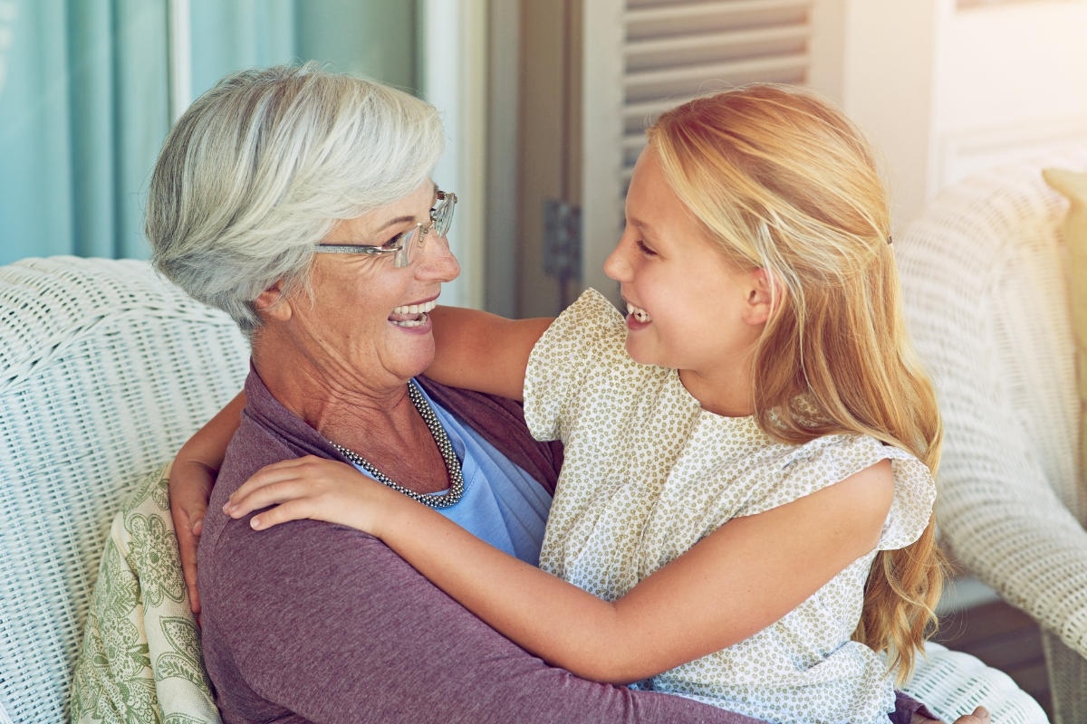 Une femme âgée souriante aux cheveux gris courts enlace une jeune fille blonde alors qu’elles sont assises ensemble à l’intérieur, partageant un moment joyeux.