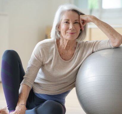 Femme senior souriante en tenue de sport, assise dans un salon, penchée sur une balle de pilates.