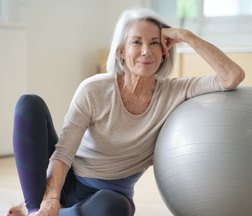 Femme senior souriante en tenue de sport, assise dans un salon, penchée sur une balle de pilates.