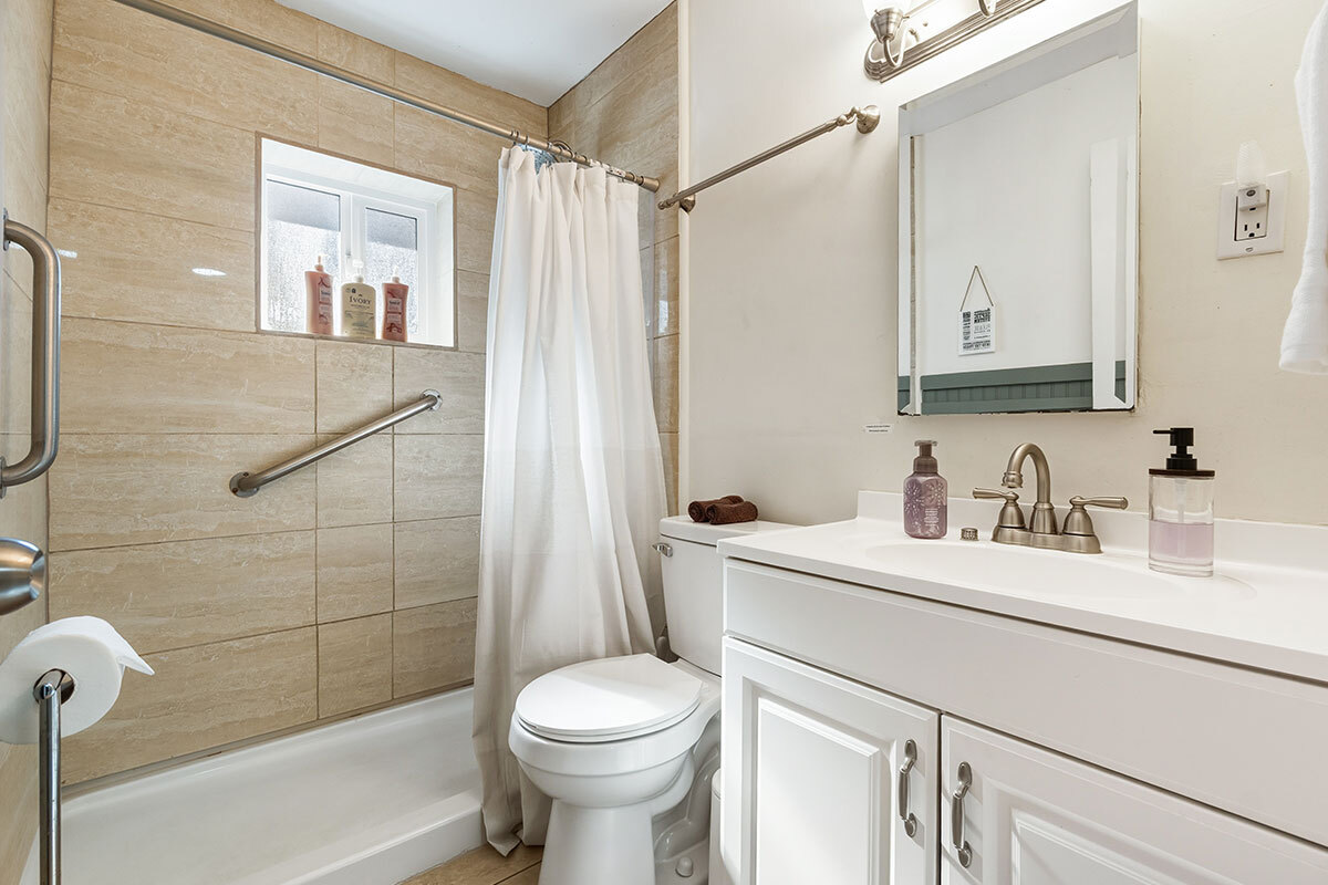 Salle de bains avec baignoire-douche équipée d'une barre d'appui, rideau blanc, meuble-vasque blanc et miroir. 