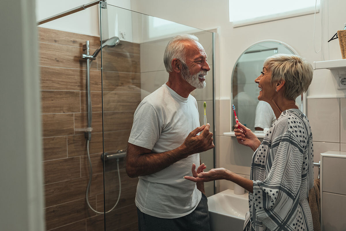 Un couple de personnes âgées se brosse les dents en riant ensemble dans une salle de bains moderne, avec douche à l'italienne en arrière-plan.