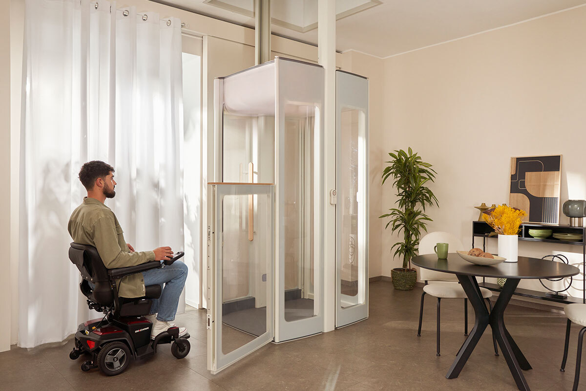 Un homme en fauteuil roulant électrique actionne la télécommande d'un ascenseur domestique vitré Uplifts S3, dans un salon lumineux. 