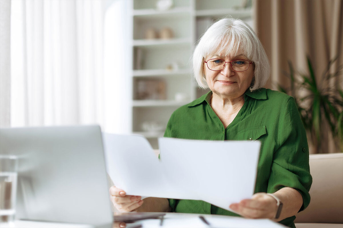 Une femme âgée aux cheveux blancs et lunettes, assise à un bureau, tenant des documents devant un ordinateur portable. 