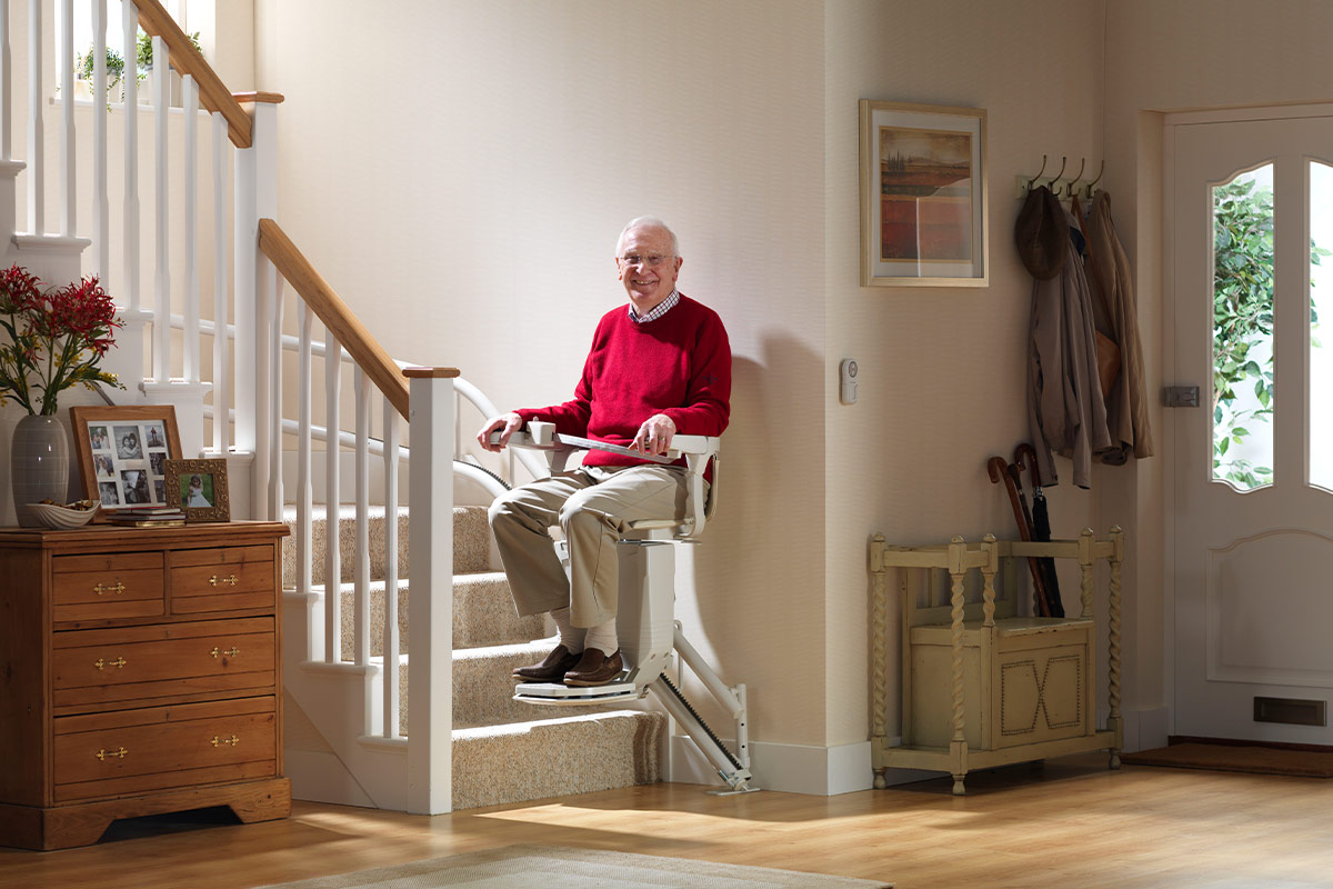 Un homme âgé souriant est assis sur un monte-escalier installé dans un escalier d'intérieur, près d'une entrée avec porte-manteaux et commode en bois.