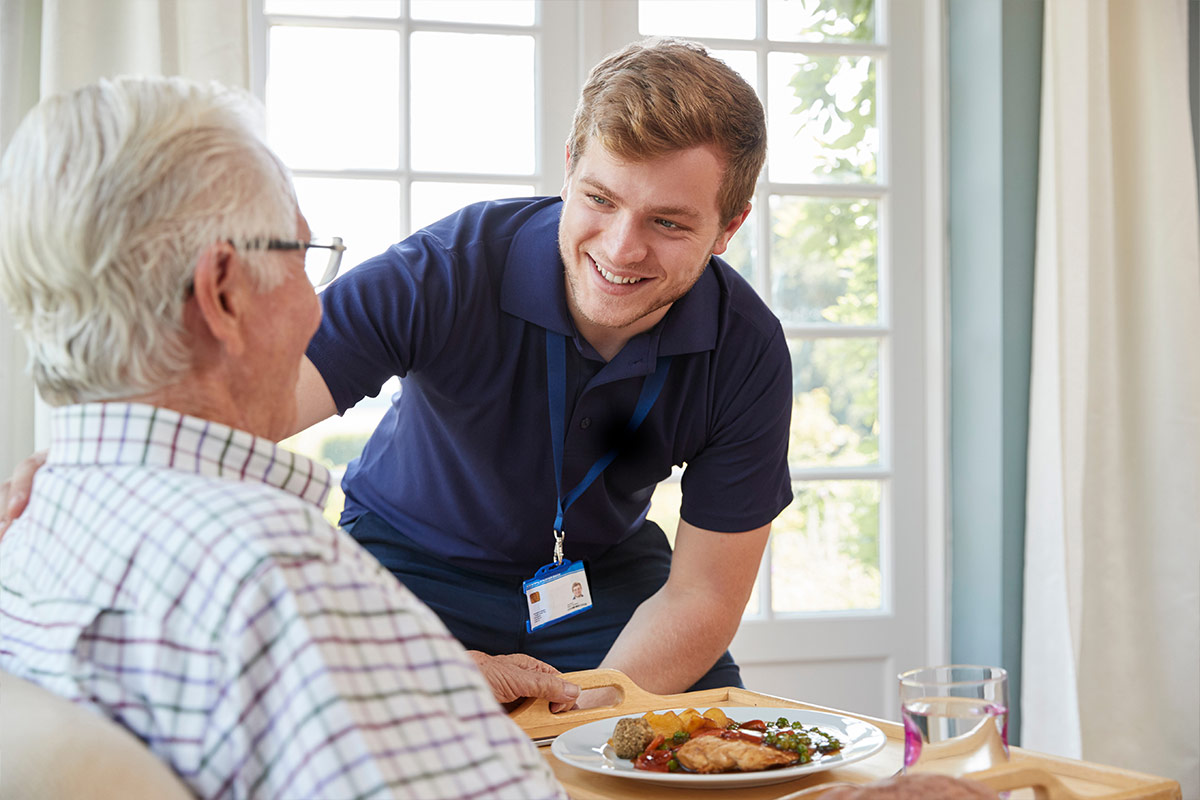 Un jeune aidant souriant apporte un plateau-repas à un homme âgé assis à table. 