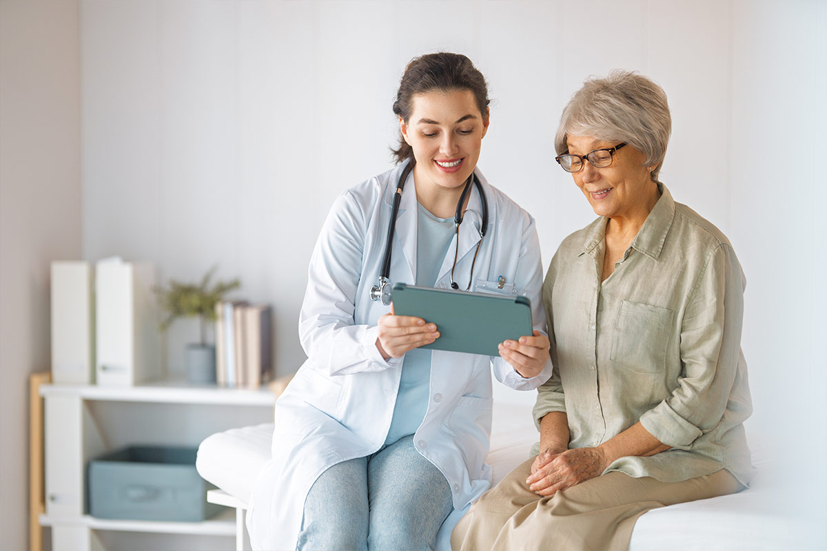 Une médecin montre des informations sur une tablette à une patiente âgée, assise à ses côtés dans un cabinet médical.