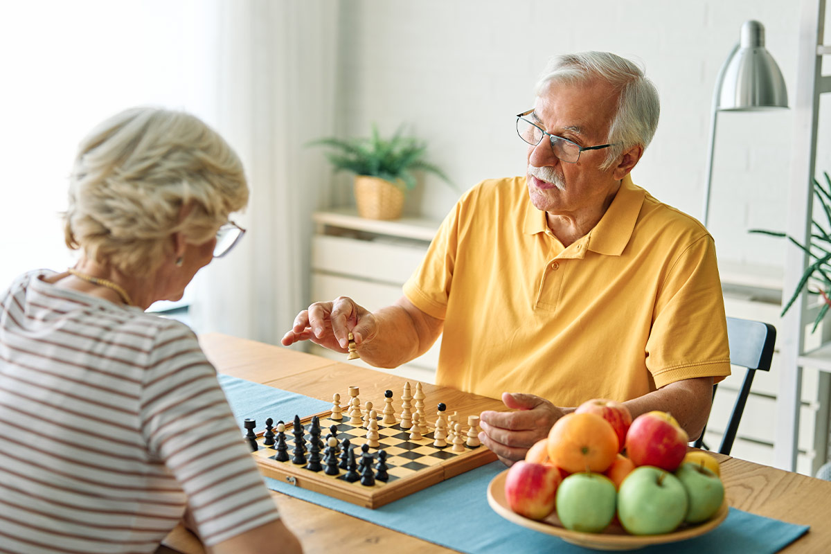 Un couple de personnes âgées joue aux échecs à table, avec un bol de fruits frais posé à côté du plateau de jeu