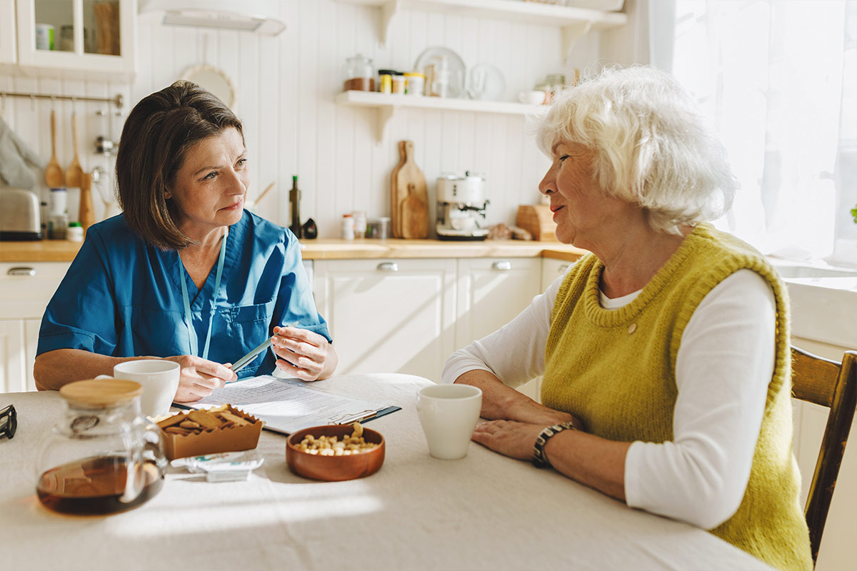 Une aide-soignante en tenue bleue discute avec une femme âgée autour d'une table, dans une cuisine lumineuse.