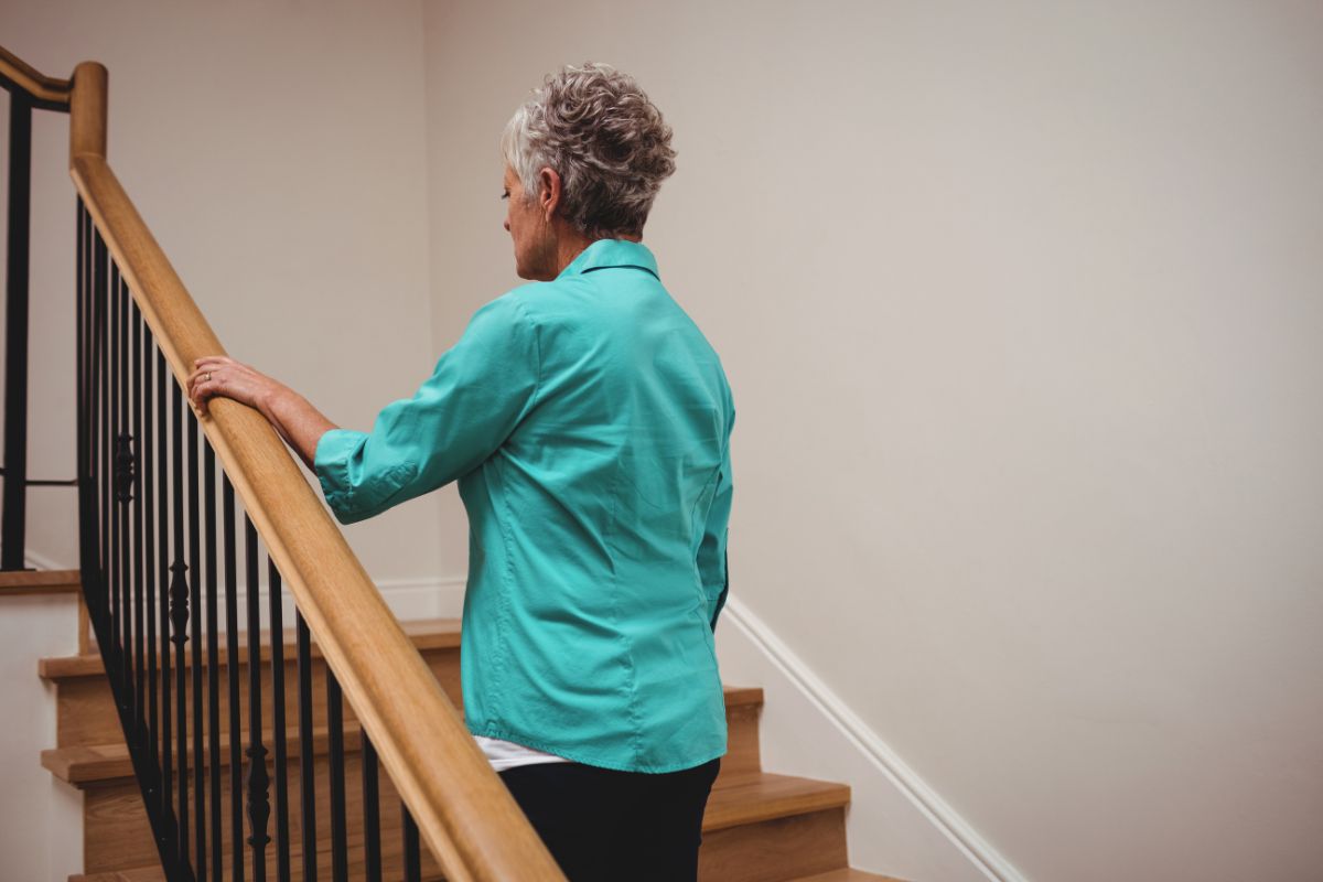 Une femme âgée aux cheveux gris descend un escalier en bois en tenant la rampe. 