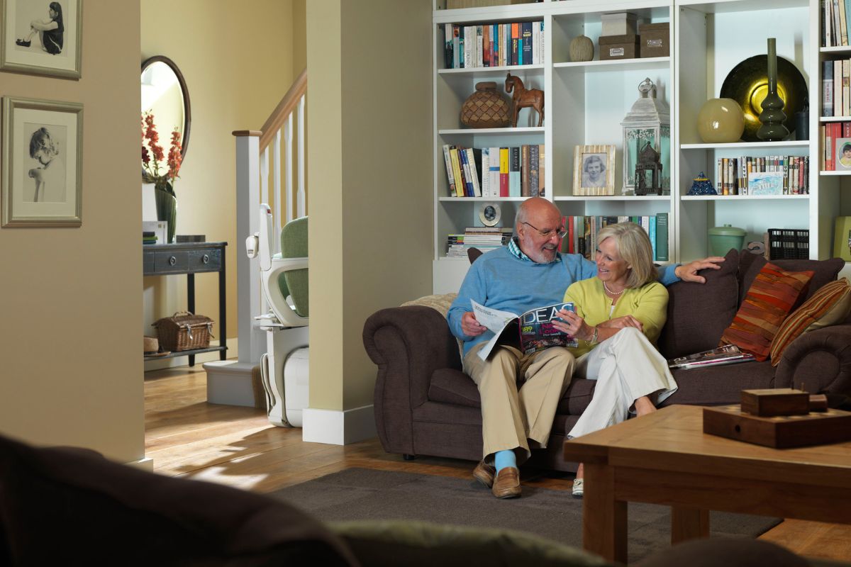Un couple de personnes âgées assis sur un canapé, lisant un magazine ensemble dans un salon avec bibliothèque et monte-escalier visible en arrière-plan. 