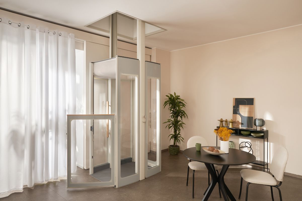 Un ascenseur domestique vitré Uplifts S3 installé dans un salon, entre un rideau blanc et une table à manger noire. 