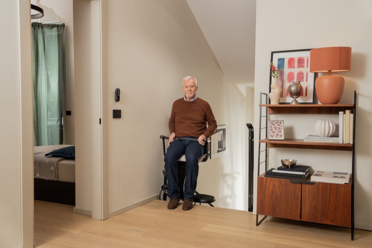 Un homme âgé souriant assis sur un monte-escalier compact, dans un intérieur moderne près d'une chambre et d'une bibliothèque en bois.