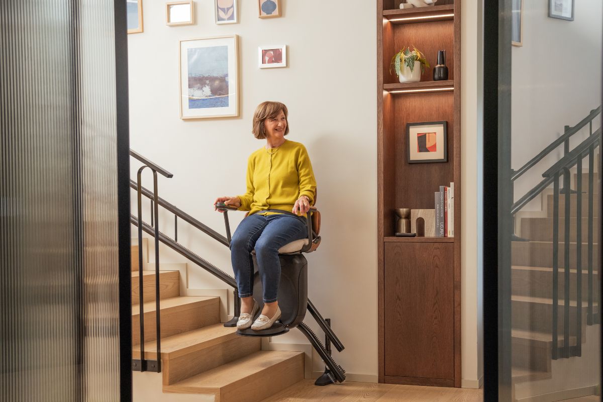 Une femme souriante en pull jaune descend un escalier en bois sur un monte-escalier Stannah Joya, vue à travers une porte vitrée, dans un intérieur moderne.
