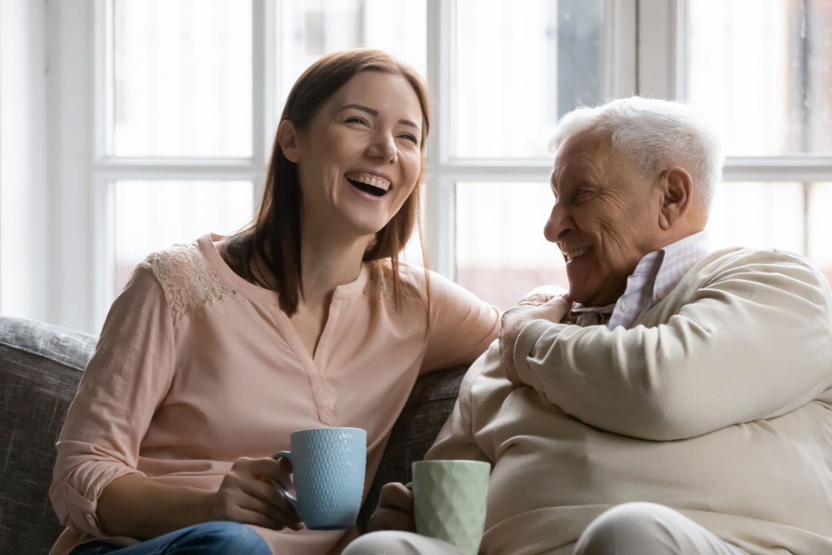  Une jeune femme rit avec un homme âgé, tous deux tenant une tasse, assis côte à côte sur un canapé.