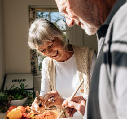 Un couple de personnes âgées cuisine ensemble dans une cuisine lumineuse, la femme souriante coupant des légumes pendant que l'homme l'assiste.