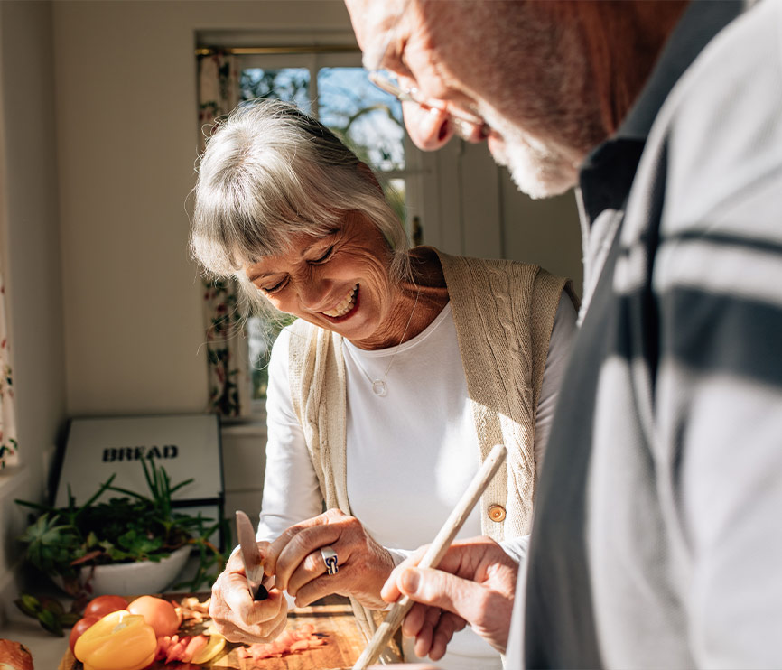 Un couple de personnes âgées cuisine ensemble dans une cuisine lumineuse, la femme souriante coupant des légumes pendant que l'homme l'assiste.
