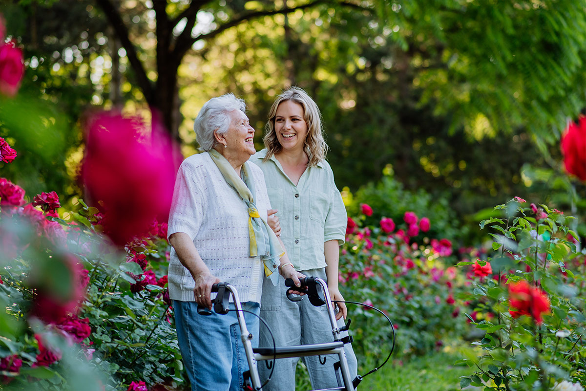 Une aidante souriante accompagne une femme âgée utilisant un déambulateur dans un jardin fleuri de roses.