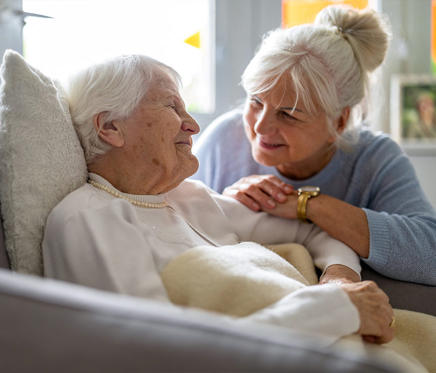Deux femmes âgées partagent un moment tendre, l'une allongée sur un canapé et l'autre penchée vers elle avec affection.