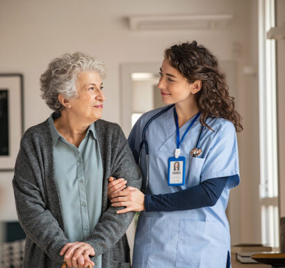 Une infirmière en tenue bleue accompagne une femme âgée souriante, la main posée sur son bras, dans un couloir intérieur.