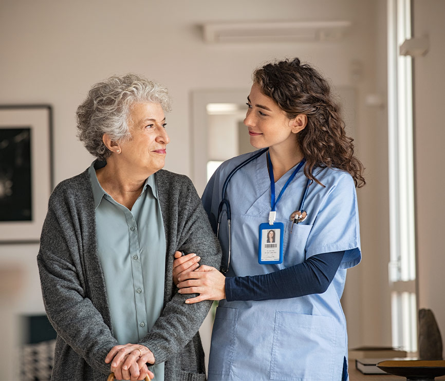 Une infirmière en tenue bleue accompagne une femme âgée souriante, la main posée sur son bras, dans un couloir intérieur.