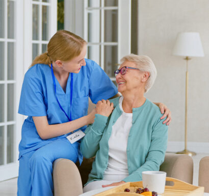 Une infirmière en tenue bleue pose la main sur l'épaule d'une femme âgée souriante, assise dans un salon près d'une baie vitrée.