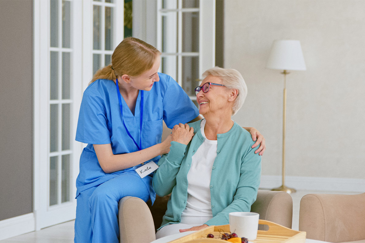 Une infirmière en tenue bleue pose la main sur l'épaule d'une femme âgée souriante, assise dans un salon près d'une baie vitrée.