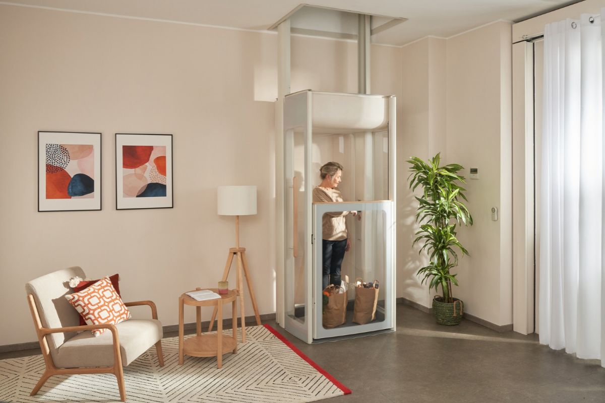 Femme senior dans un ascenseur de maison Stannah vitré avec deux sacs de courses, dans un salon lumineux et moderne