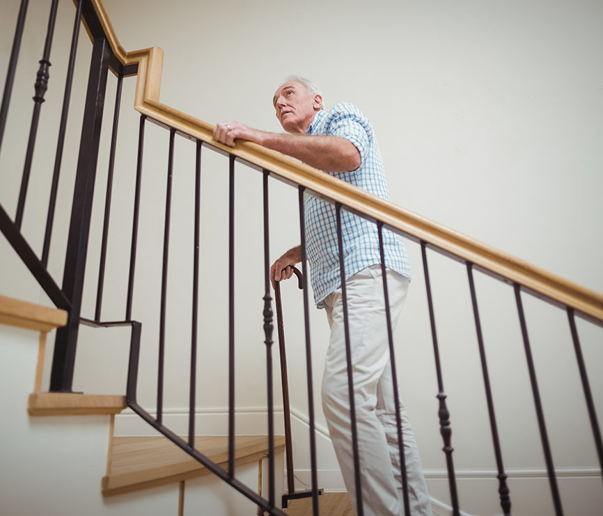 Homme senior avec une canne qui monte un escalier en s'appuyant sur la rampe, le regard tourné vers l'étage 