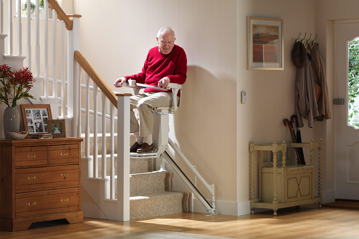 Homme senior souriant assis sur un monte-escalier Stannah blanc, installé sur un escalier droit dans une entrée chaleureuse