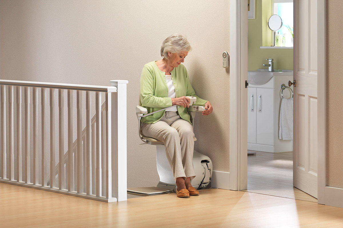 Femme senior assise sur un monte-escalier Stannah qui attache sa ceinture en haut de l'escalier, près de la salle de bain